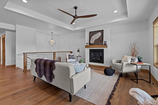 living area featuring a glass covered fireplace, recessed lighting, a ceiling fan, wood finished floors, and a tray ceiling