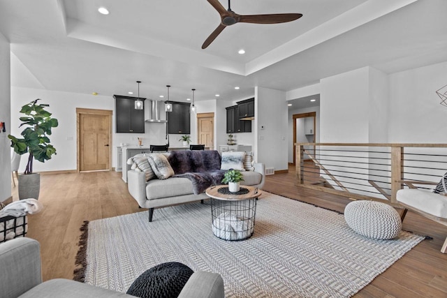 living room with ceiling fan, a raised ceiling, baseboards, light wood finished floors, and recessed lighting