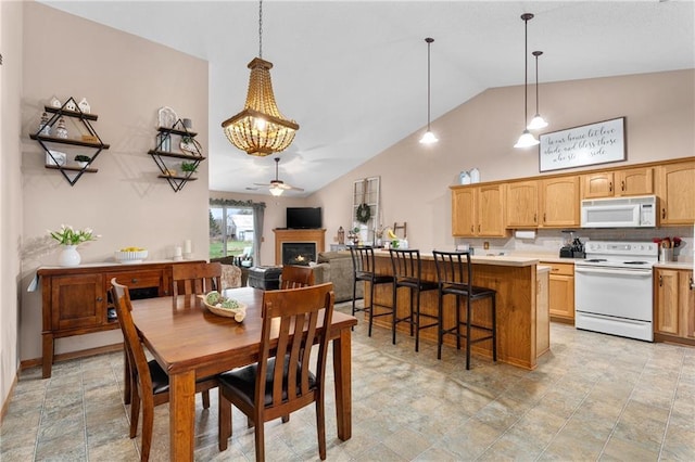 dining area with ceiling fan with notable chandelier, stone finish floor, a lit fireplace, and high vaulted ceiling