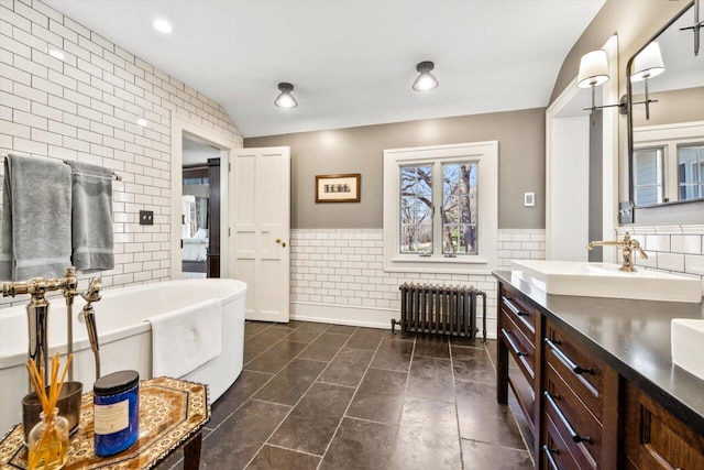 full bath featuring a sink, double vanity, radiator, a freestanding tub, and tile walls