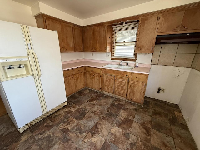 kitchen featuring a sink, white refrigerator with ice dispenser, light countertops, brown cabinetry, and stone finish flooring