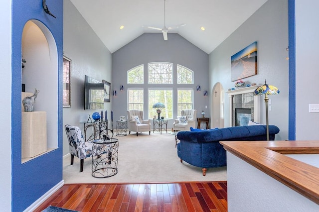 living area with a ceiling fan, a tiled fireplace, arched walkways, high vaulted ceiling, and wood-type flooring