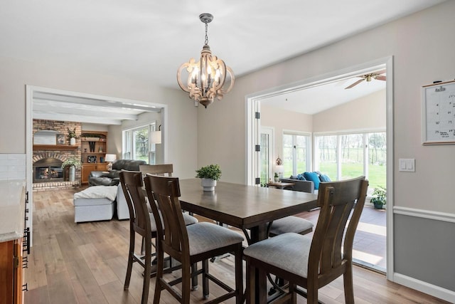 dining room with a fireplace, vaulted ceiling, light wood-type flooring, baseboards, and ceiling fan with notable chandelier