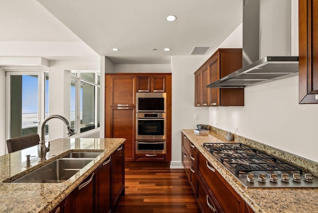 kitchen with stainless steel gas cooktop, wall chimney exhaust hood, light stone counters, a sink, and dark wood finished floors