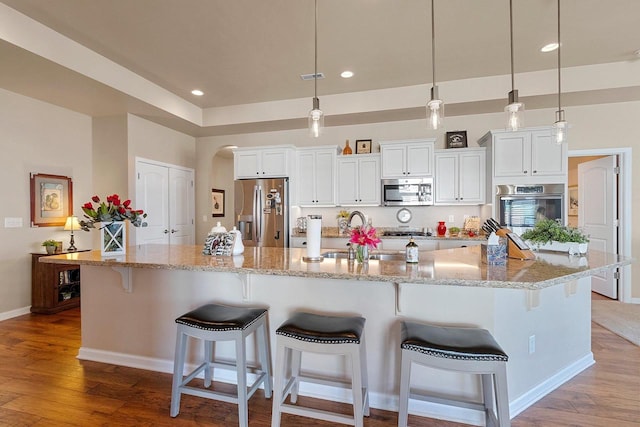 kitchen featuring appliances with stainless steel finishes, arched walkways, a large island with sink, wood finished floors, and white cabinetry