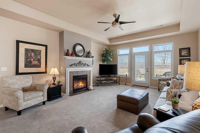 carpeted living area with visible vents, a fireplace, baseboards, a tray ceiling, and a ceiling fan