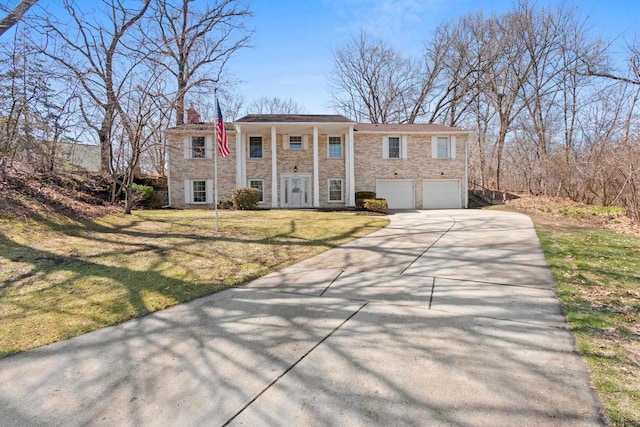 greek revival inspired property with a front lawn, stone siding, a chimney, an attached garage, and driveway