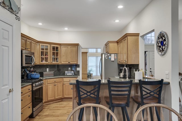 kitchen with appliances with stainless steel finishes, light wood finished floors, a peninsula, decorative backsplash, and glass insert cabinets