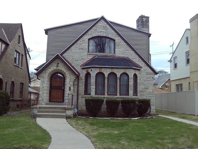 tudor home with a high end roof, a chimney, fence, a front lawn, and stone siding