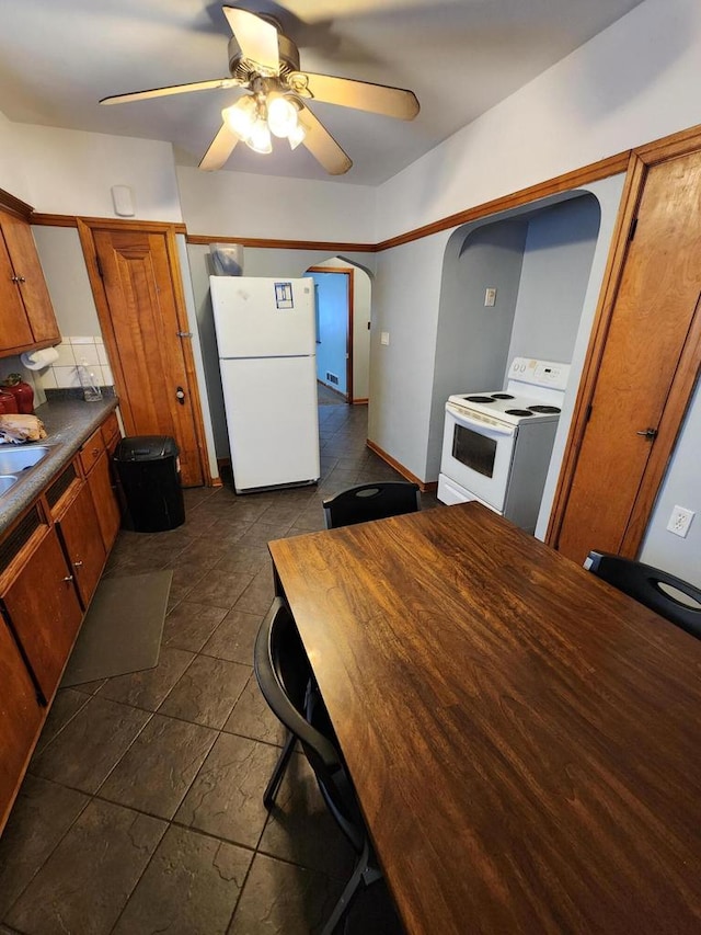 kitchen featuring dark countertops, white appliances, arched walkways, brown cabinetry, and a ceiling fan