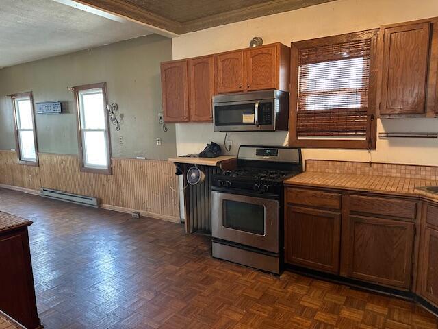 kitchen with baseboard heating, wainscoting, wood walls, brown cabinets, and stainless steel appliances