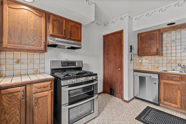 kitchen with tile countertops, under cabinet range hood, a sink, appliances with stainless steel finishes, and backsplash