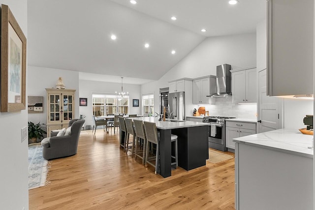 kitchen with appliances with stainless steel finishes, a kitchen breakfast bar, an island with sink, wall chimney exhaust hood, and light wood-type flooring