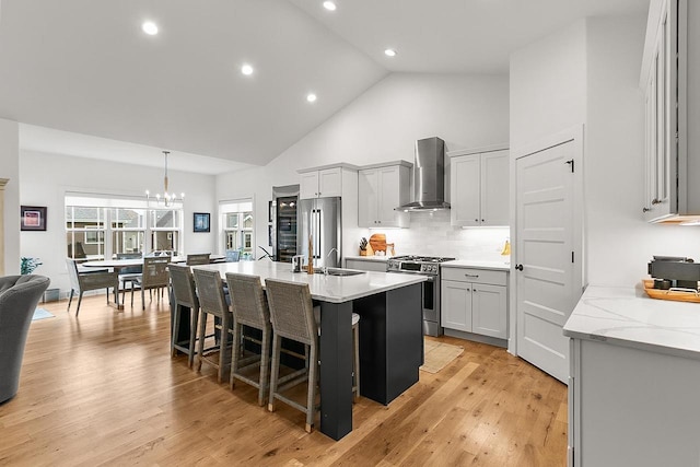 kitchen with wall chimney range hood, high vaulted ceiling, a breakfast bar, stainless steel appliances, and an island with sink