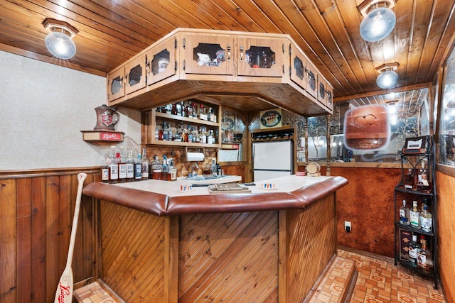 bar featuring freestanding refrigerator, brick floor, a wainscoted wall, wooden ceiling, and a bar