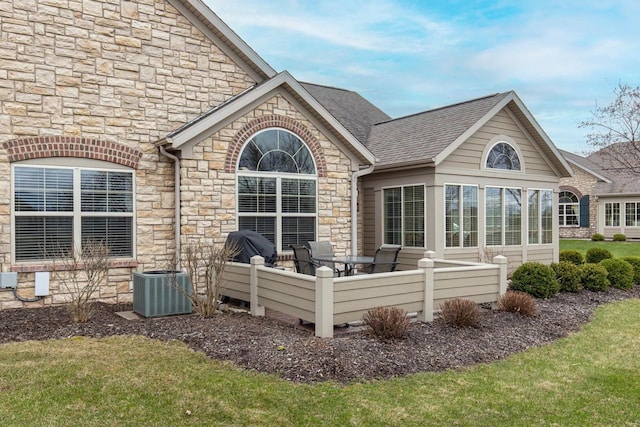 back of property featuring a lawn, central AC unit, stone siding, and a shingled roof