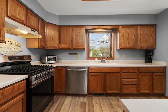 kitchen with brown cabinetry, a sink, light countertops, under cabinet range hood, and stainless steel appliances