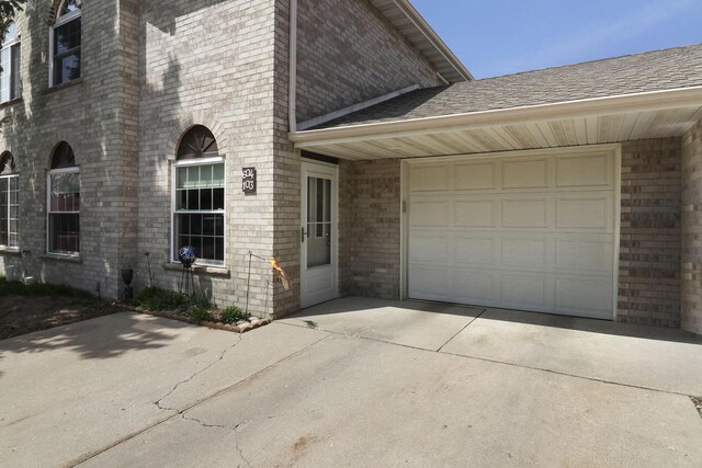 view of exterior entry featuring a shingled roof, an attached garage, concrete driveway, and brick siding
