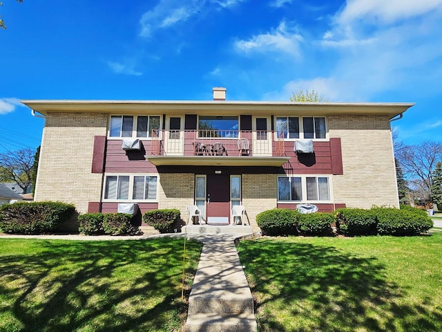 view of property with brick siding, a chimney, a balcony, and a front yard