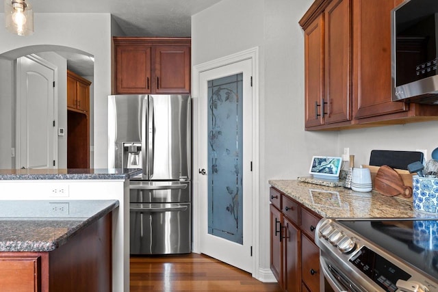 kitchen with dark wood-style flooring, dark stone counters, brown cabinets, arched walkways, and stainless steel appliances