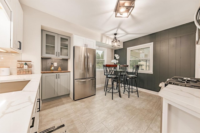 kitchen with white cabinets, light stone counters, freestanding refrigerator, backsplash, and glass insert cabinets