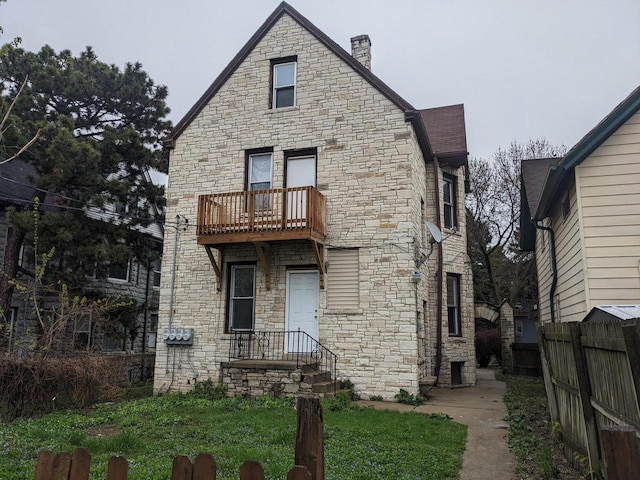 back of house featuring fence, a chimney, a balcony, and stone siding