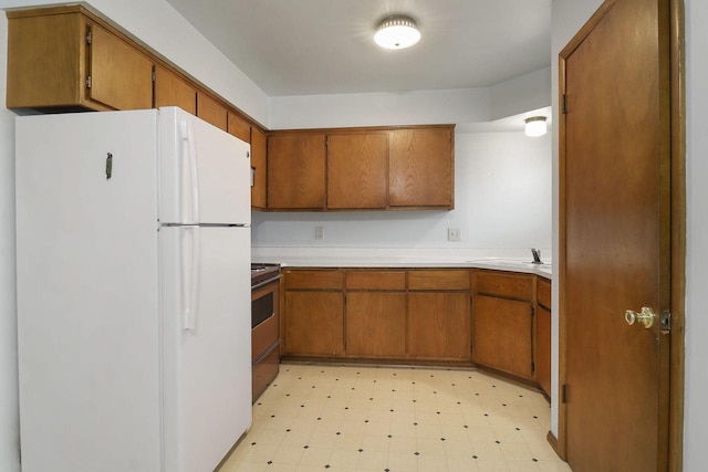kitchen with brown cabinetry, light countertops, freestanding refrigerator, electric stove, and light floors