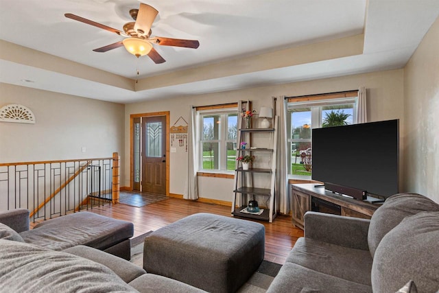 living area featuring a raised ceiling, ceiling fan, baseboards, and wood finished floors
