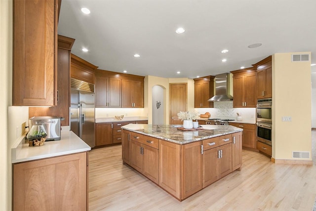 kitchen with wall chimney exhaust hood, appliances with stainless steel finishes, a kitchen island, light wood-type flooring, and arched walkways