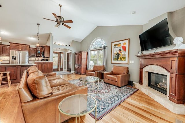 living area featuring a ceiling fan, high vaulted ceiling, a glass covered fireplace, and light wood-style flooring