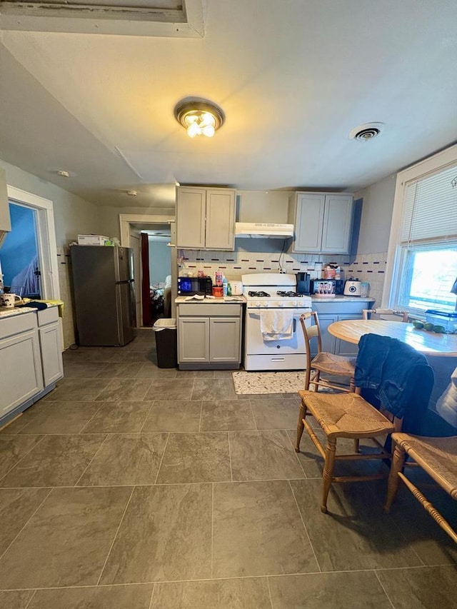 kitchen with freestanding refrigerator, white gas stove, decorative backsplash, under cabinet range hood, and visible vents