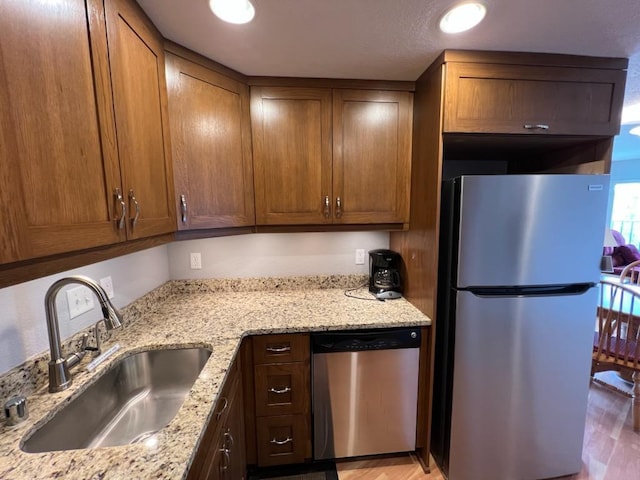 kitchen with brown cabinetry, appliances with stainless steel finishes, light stone countertops, a sink, and light wood-style floors