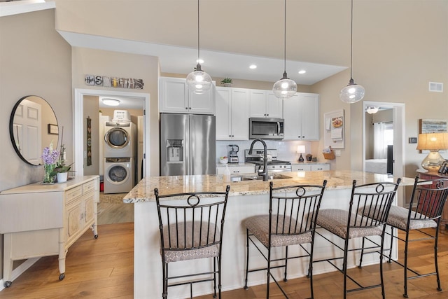 kitchen with stainless steel appliances, stacked washer and dryer, light wood-type flooring, light stone counters, and an island with sink