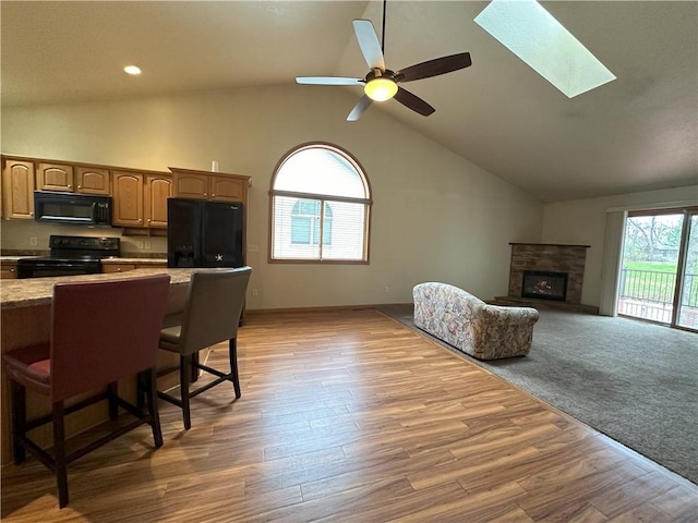 interior space with light wood-style floors, high vaulted ceiling, a stone fireplace, a ceiling fan, and a skylight
