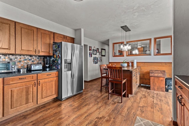 kitchen featuring brown cabinetry, tasteful backsplash, stainless steel refrigerator with ice dispenser, dark wood finished floors, and dark countertops
