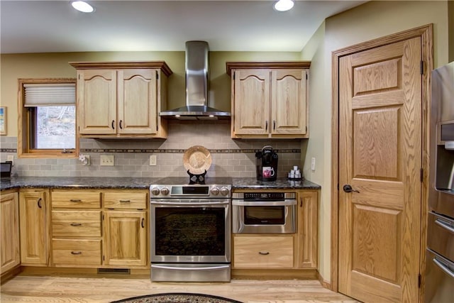 kitchen featuring tasteful backsplash, light wood-type flooring, wall chimney exhaust hood, light brown cabinetry, and appliances with stainless steel finishes