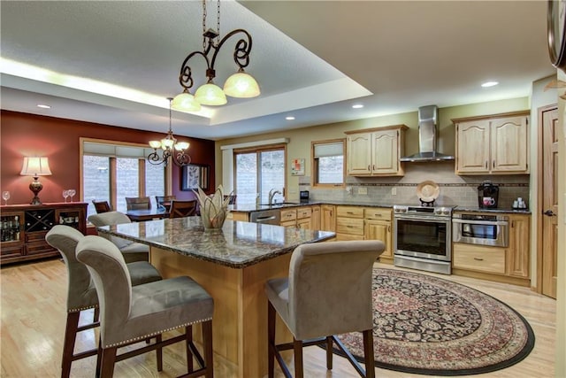 kitchen with light brown cabinetry, a breakfast bar area, an inviting chandelier, wall chimney exhaust hood, and stainless steel appliances