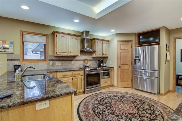 kitchen featuring a sink, wall chimney range hood, dark stone countertops, light wood-type flooring, and appliances with stainless steel finishes