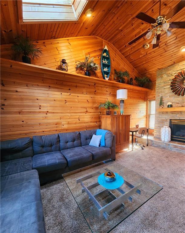 carpeted living room featuring a skylight, wood walls, ceiling fan, wood ceiling, and a stone fireplace