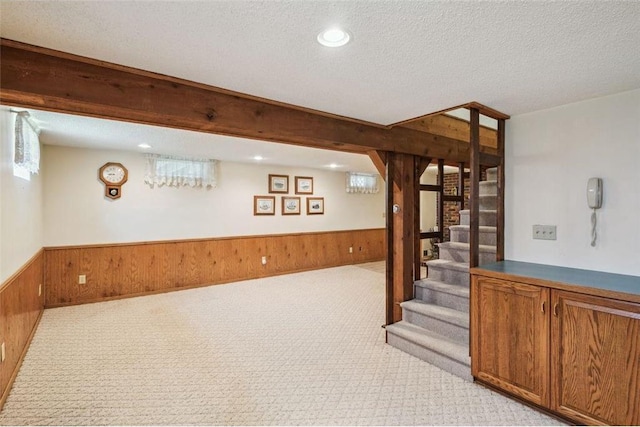 basement featuring stairs, light colored carpet, a textured ceiling, wooden walls, and wainscoting