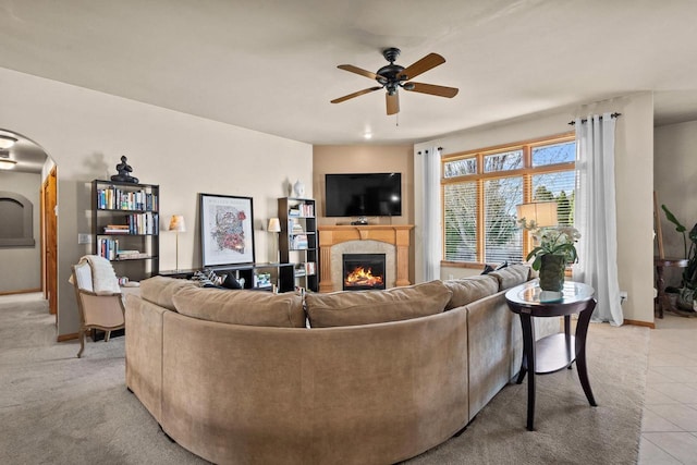 living room featuring a ceiling fan, arched walkways, a warm lit fireplace, light tile patterned flooring, and baseboards
