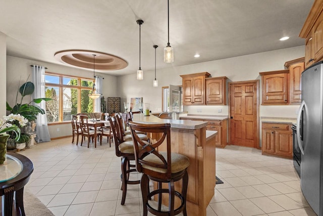 kitchen with freestanding refrigerator, a kitchen breakfast bar, brown cabinetry, light tile patterned floors, and recessed lighting