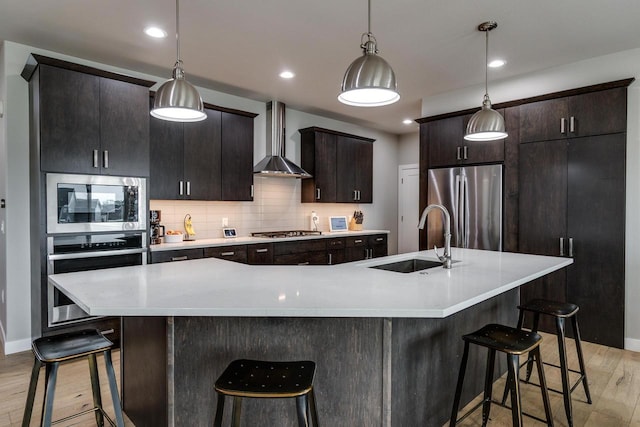 kitchen with a sink, appliances with stainless steel finishes, wall chimney range hood, light wood-type flooring, and light countertops