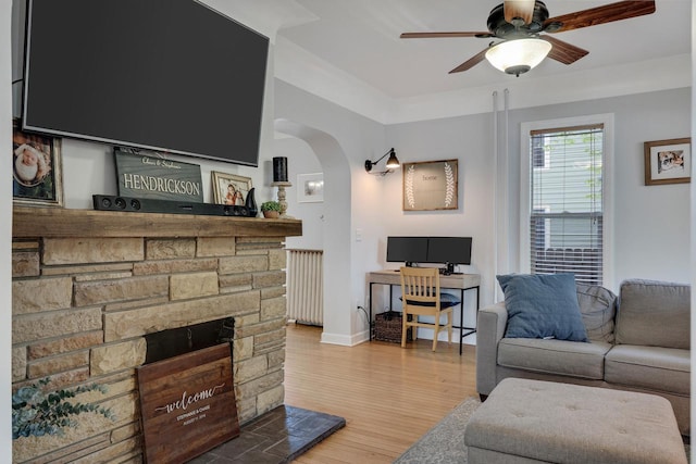 living area with radiator heating unit, arched walkways, a ceiling fan, wood finished floors, and a stone fireplace