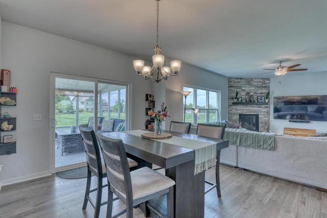 dining area featuring light wood-style flooring, a stone fireplace, ceiling fan, a chandelier, and baseboards