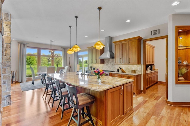 7216 W Fox Haven Ct, Franklin, WI 53132 - kitchen featuring wall chimney exhaust hood, light wood-style flooring, decorative backsplash, light stone counters, and a center island with sink