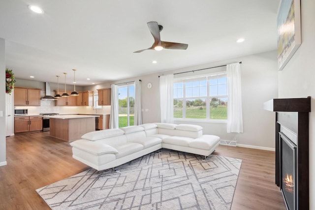 living area featuring a lit fireplace, light wood-style flooring, a ceiling fan, baseboards, and recessed lighting