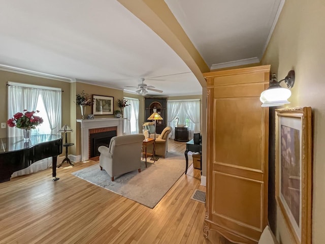 living area featuring ornamental molding, a brick fireplace, light wood-type flooring, ceiling fan, and arched walkways