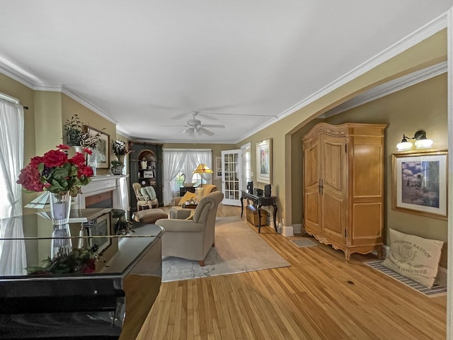 living room featuring light wood finished floors, a ceiling fan, a fireplace, arched walkways, and crown molding