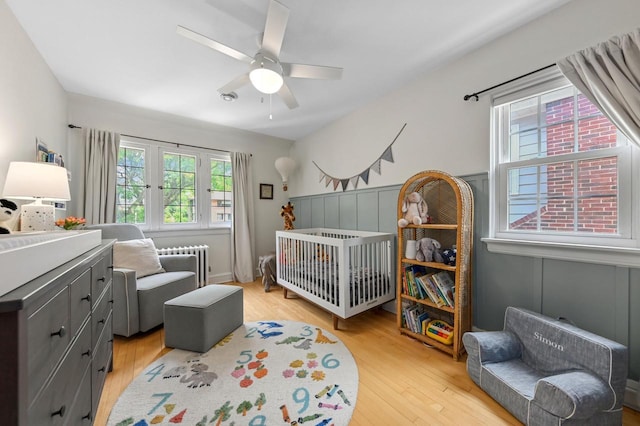 6223 W Wisconsin Ave, Wauwatosa, WI 53213 - bedroom featuring a crib, radiator heating unit, light wood finished floors, a wainscoted wall, and ceiling fan
